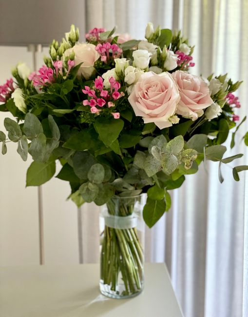 A colorful floral arrangement in a clear glass vase, featuring soft pink roses, white roses, and delicate pink and white filler flowers, accented with fresh green foliage including eucalyptus leaves. 