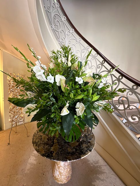 A large, elegant floral arrangement featuring white calla lilies, anthuriums, and various green foliage placed in a tall, black ceramic vase. The bouquet is crafted with a mix of lush leaves and delic