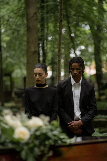 A young woman and man stand in a lush, green outdoor setting surrounded by tall trees, participating in a floral memorial or funeral service. The woman has dark hair pulled back and wears a black long