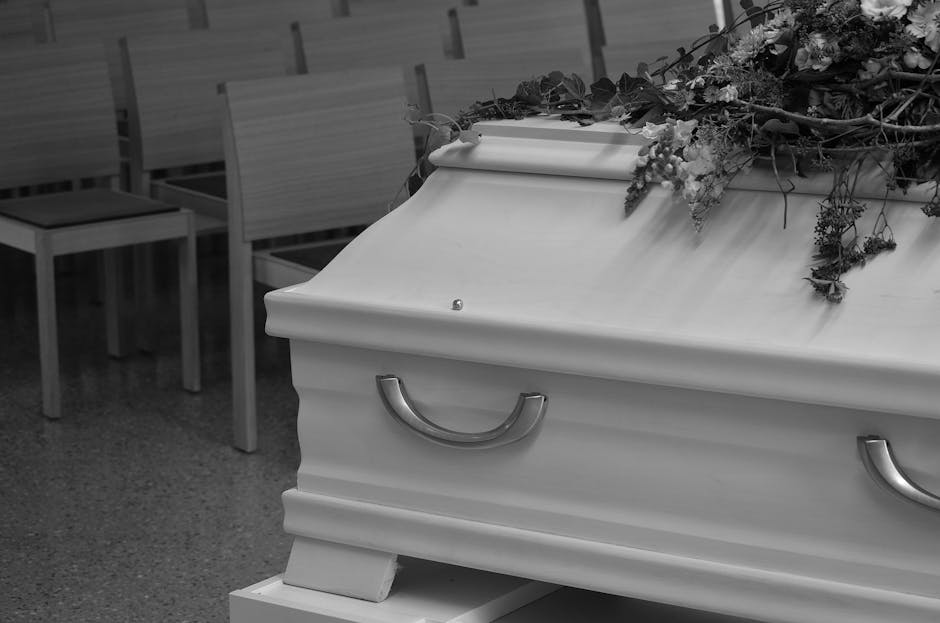 A white coffin with silver handles, topped with a floral arrangement consisting of various flowers and greenery, is placed on a stand inside a funeral home. The flowers include roses, lilies, and othe