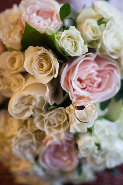 A bouquet of fresh roses arranged with a mix of white and soft pink blooms, featuring tightly curled petals and some partially opened flowers, complemented by green leaves. The bouquet is wrapped in l