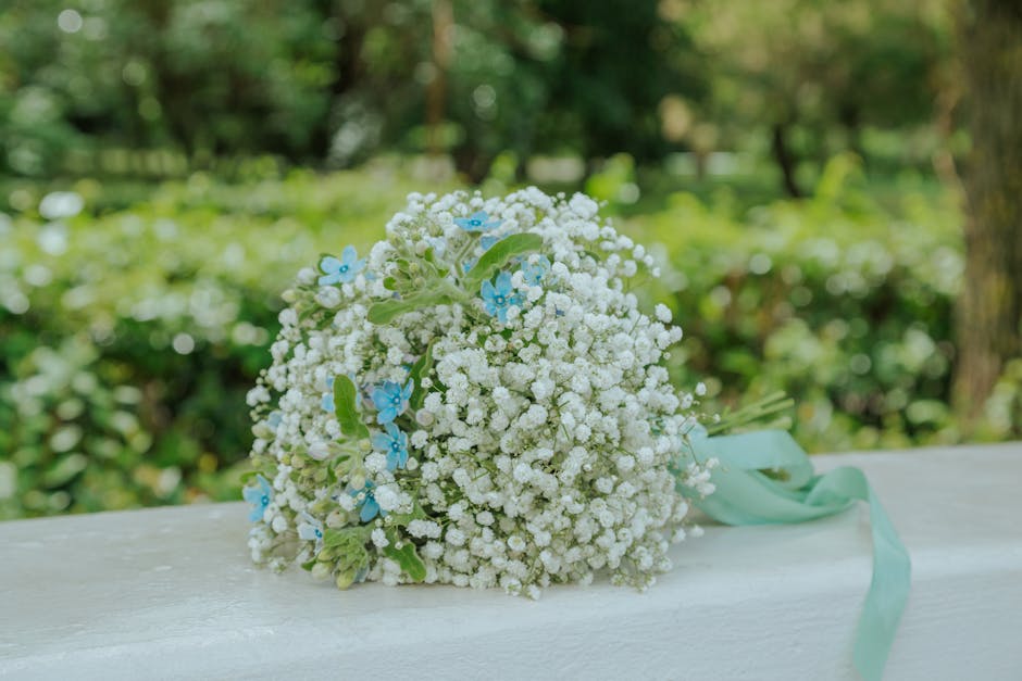 A delicate floral bouquet featuring small white Gypsophila flowers with light blue accent flowers and green foliage, wrapped with a pale mint green ribbon. The arrangement is placed on a white surface